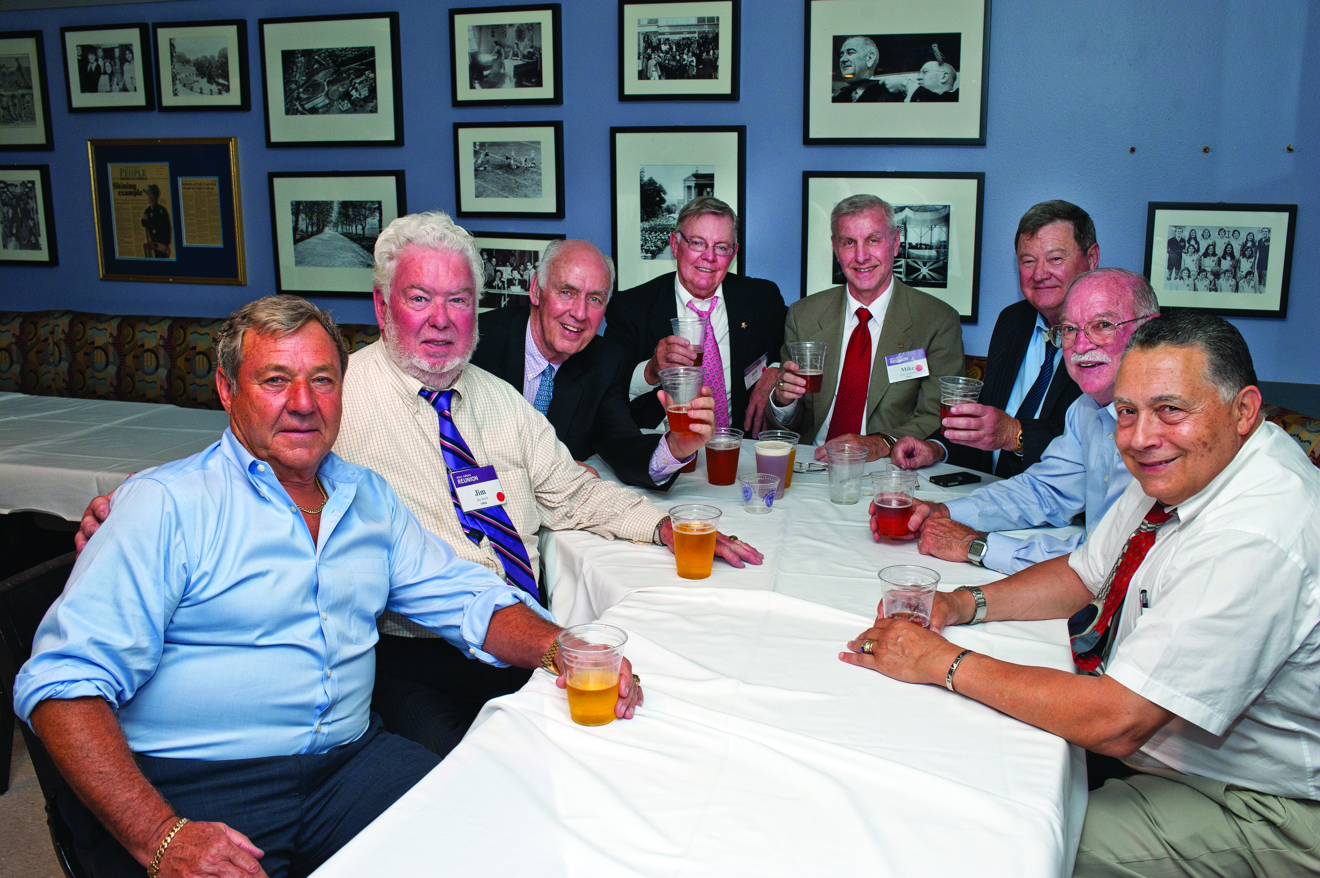 Eight men in suites and ties sit around a table covered in a tablecloth with photos on the wall in the background