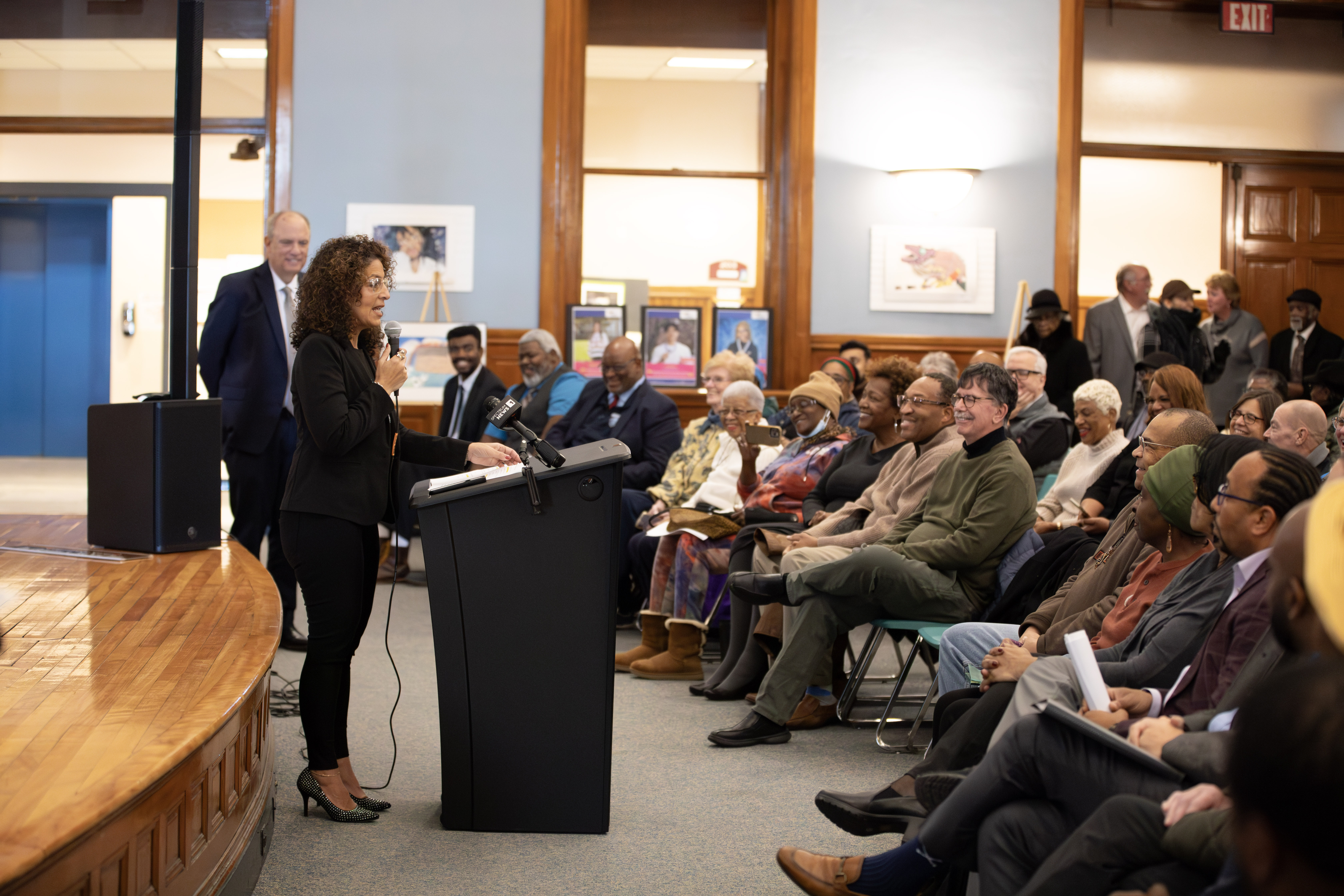 A woman stands at the front of a room with a microphone addressing a group of people.