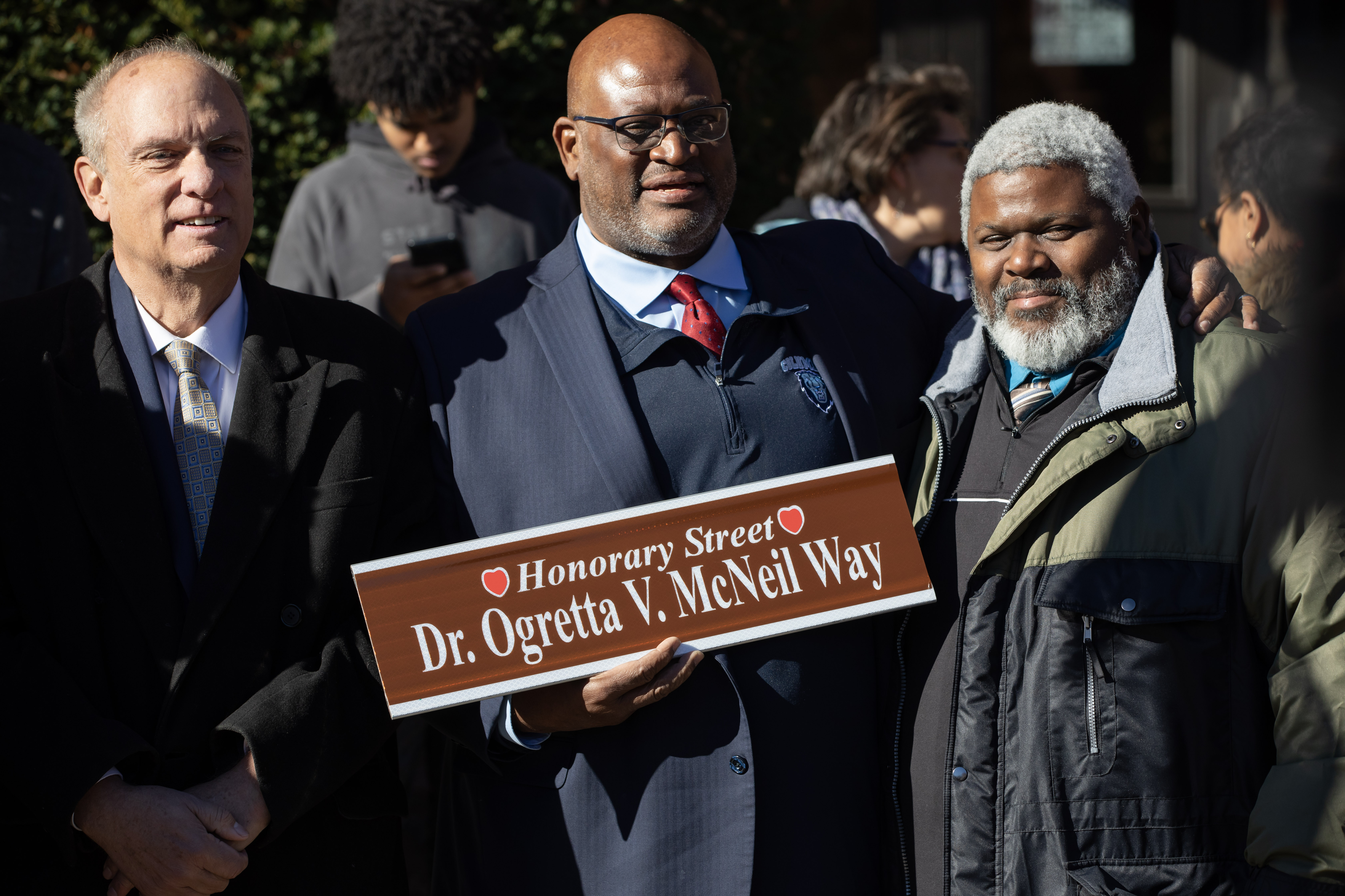 Three men stand together with the one in the middle holding a newly released street sign.