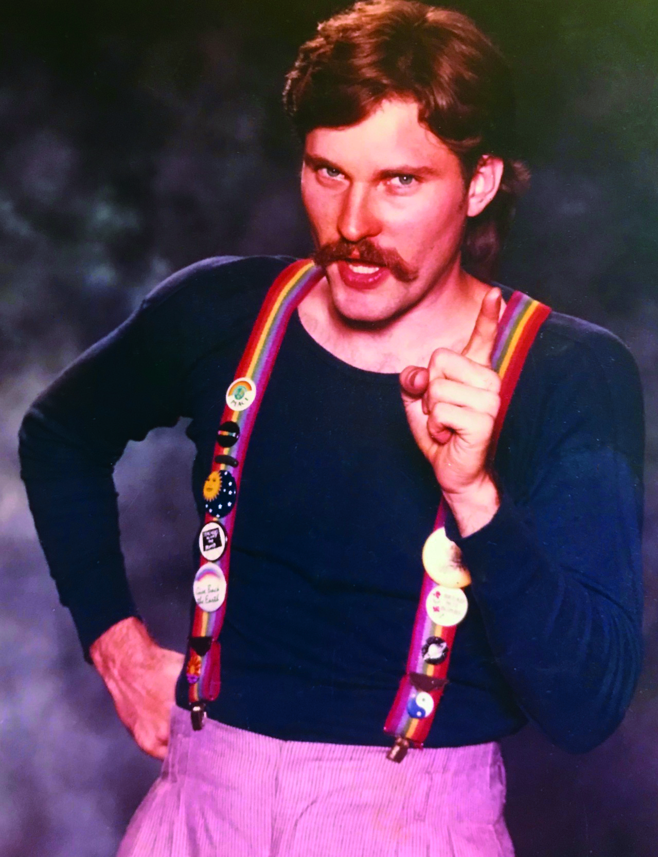 Man with moustache and rainbow suspenders points at the camera