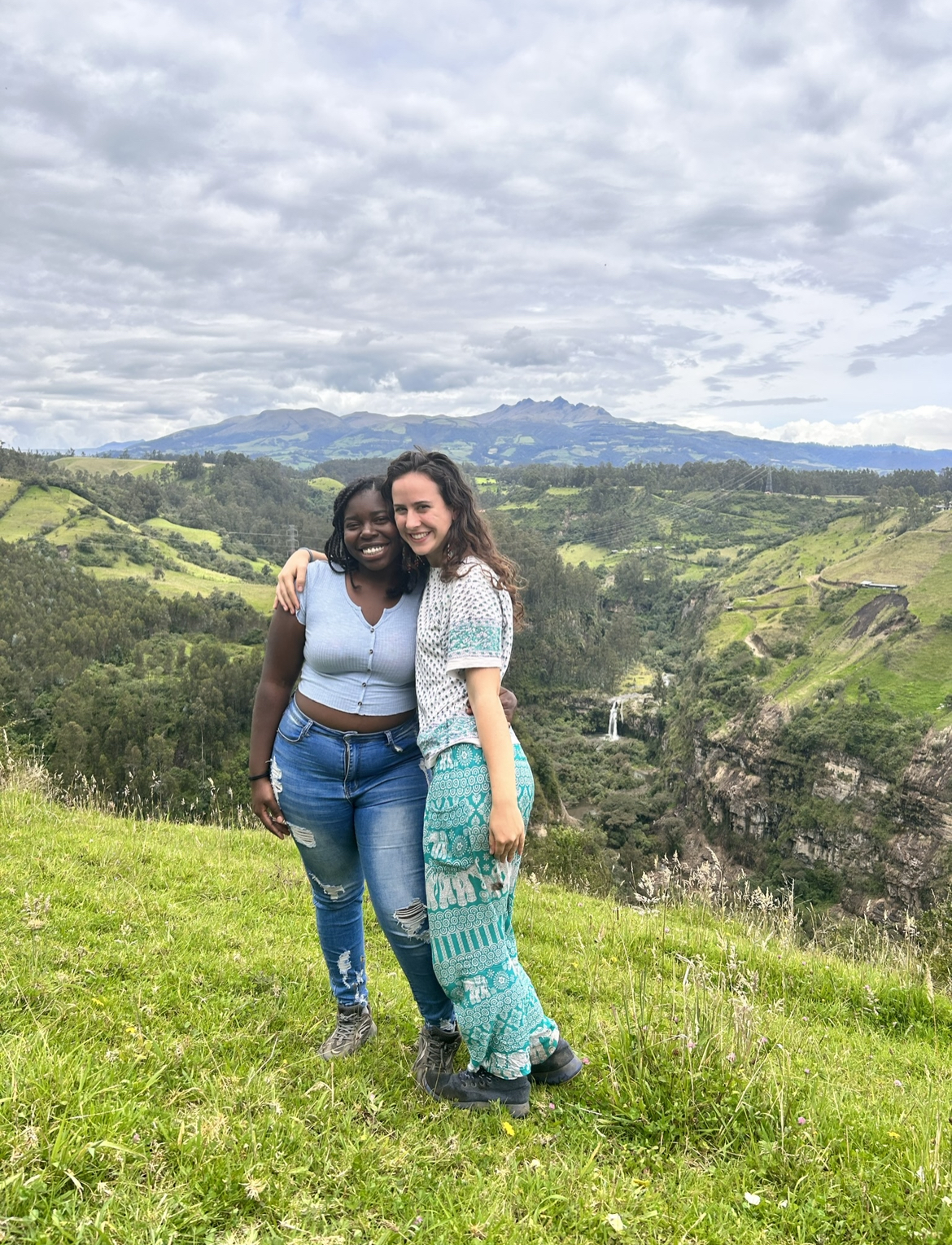 Two young woman stand next to each other in a field overlooking a valley and a mountain in the background