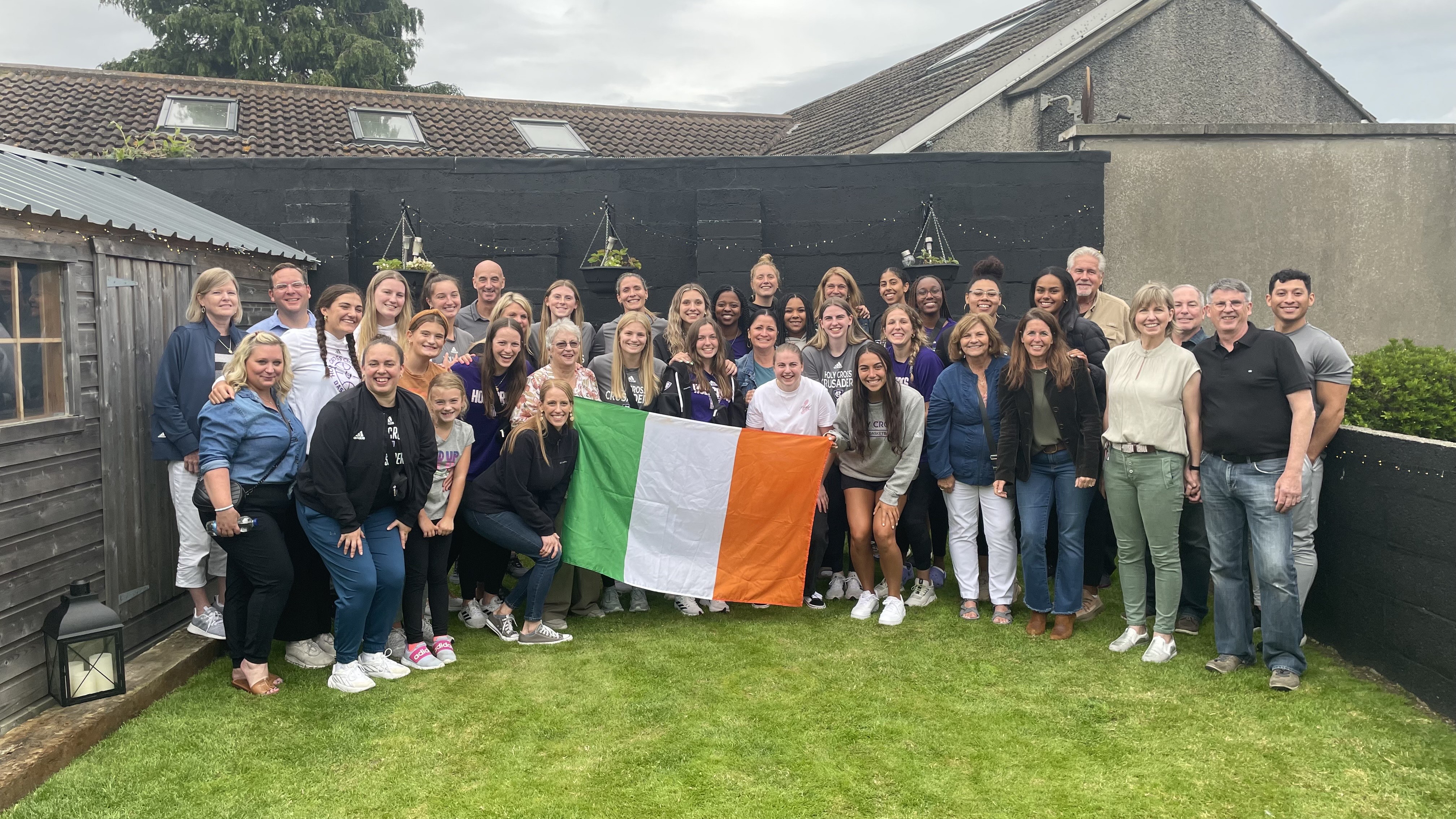 A large group of people gather in a yard holding the flag of Ireland in the center