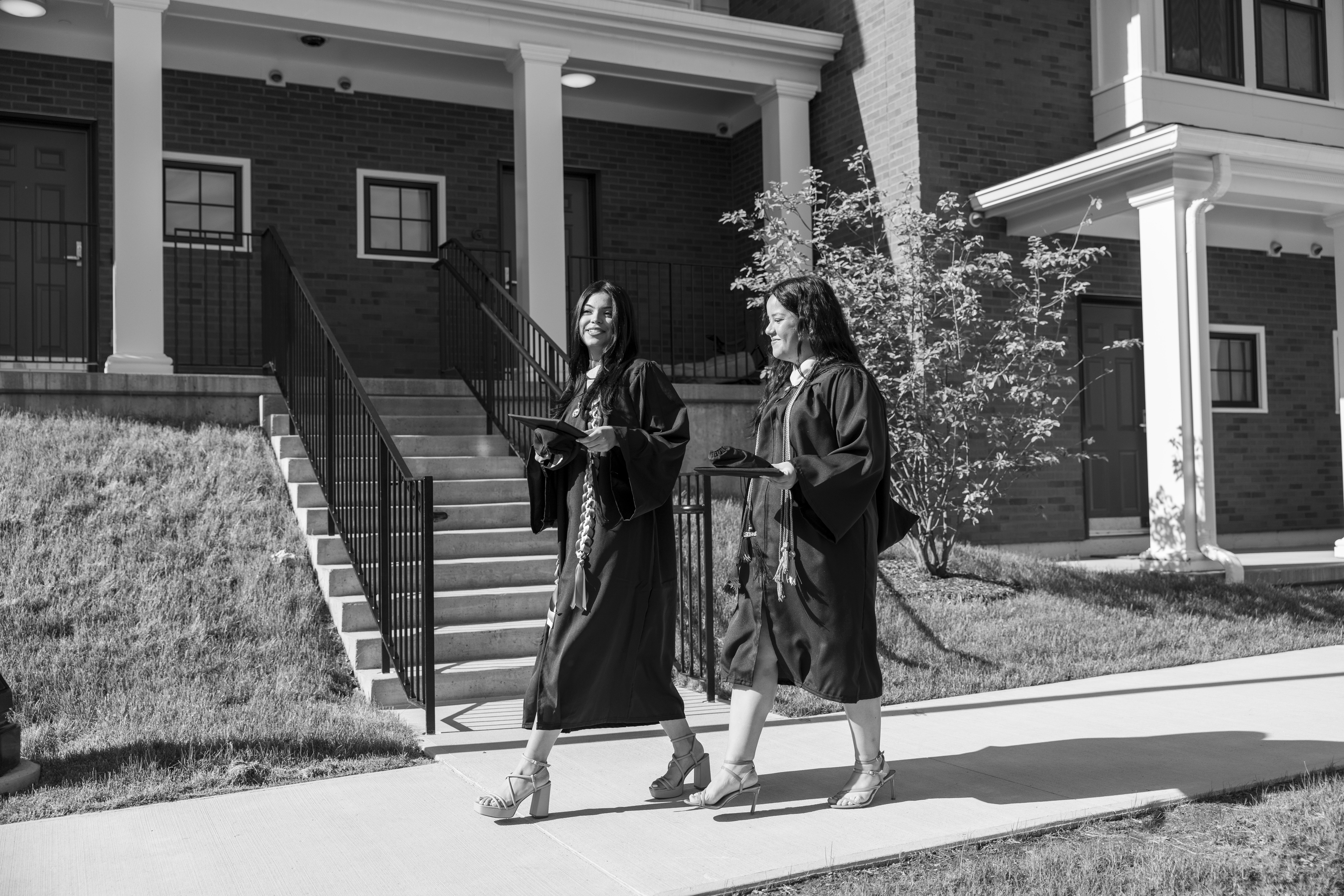Two graduates walk down the sidewalk with a townhouse behind them.