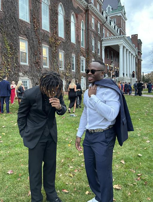 Two young men in formal suits standing in a lawn outside a brick building covered in ivy