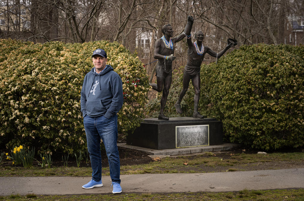 A man in a cap stands in front of a statue of two marathon runners