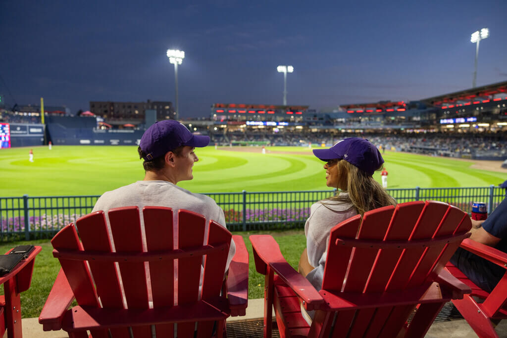 Two students sitting in red chairs at a baseball game