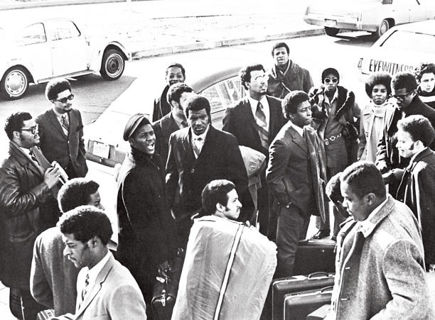 A group of mostly men gathered outside on a road by parked cars