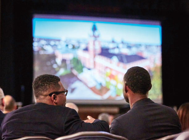 The back of two heads in front of a projector screen showing a blurred building
