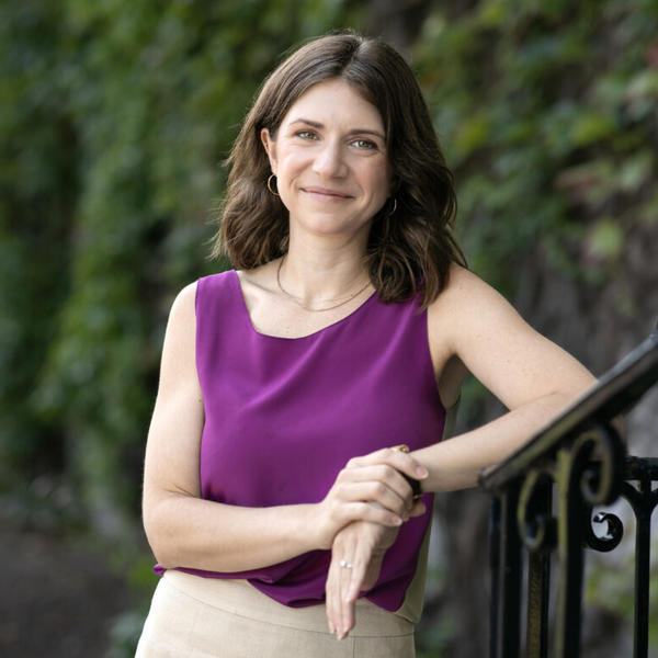 Woman dressed in purple shirt and tan skirt leans against a stair railing outside.